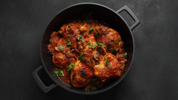 A close-up view of a pot filled with chicken thighs cooked in a savory tomato sauce, garnished with mushrooms and fresh herbs.