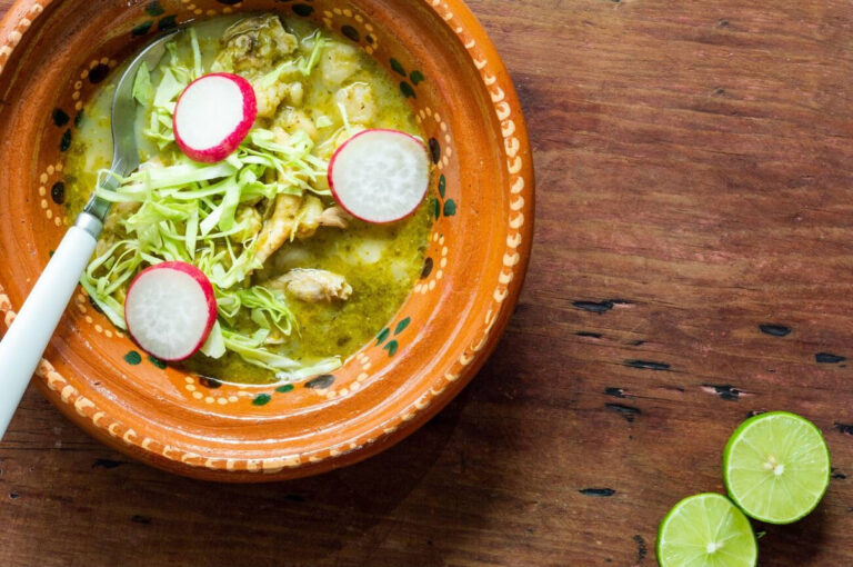 A bowl of warm soup with green herbs, pieces of chicken, and garnished with sliced radishes, placed on a wooden surface next to two lime halves.