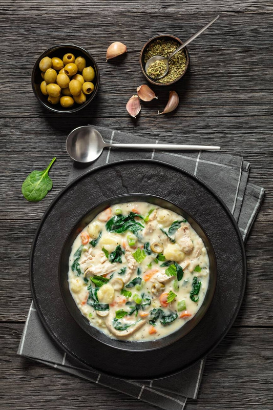 A bowl of creamy chicken and spinach soup garnished with herbs, surrounded by green olives and garlic on a wooden surface.