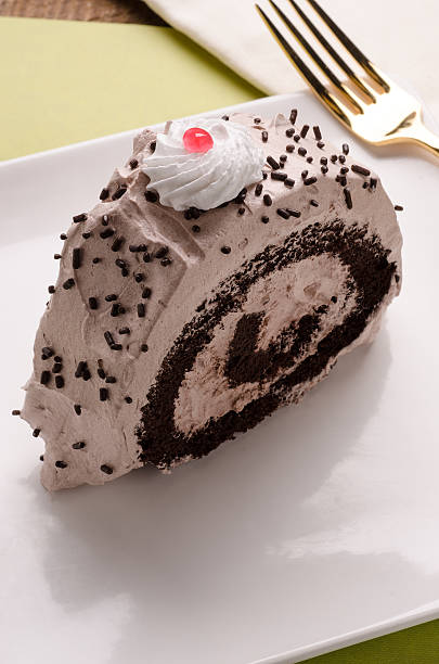 A slice of chocolate roll cake topped with whipped cream and a cherry, garnished with chocolate sprinkles, on a white plate.
