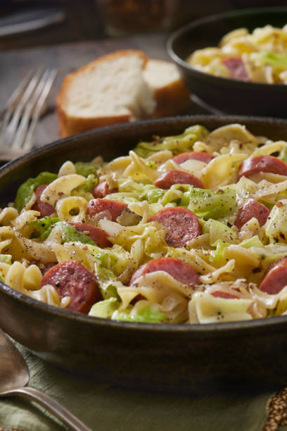 A delicious bowl of pasta with sausage and vegetables, served with slices of bread in the background.