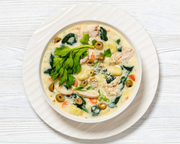 A creamy chicken soup with vegetables and olives garnished with fresh herbs in a white bowl on a wooden surface.