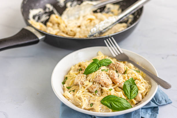 A bowl of creamy chicken fettuccine topped with fresh basil leaves, with a frying pan in the background.