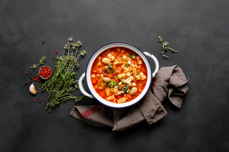 A bowl of hearty soup with macaroni, vegetables, and meat on a rustic wooden table, accompanied by fresh lettuce and a carrot on a cutting board.