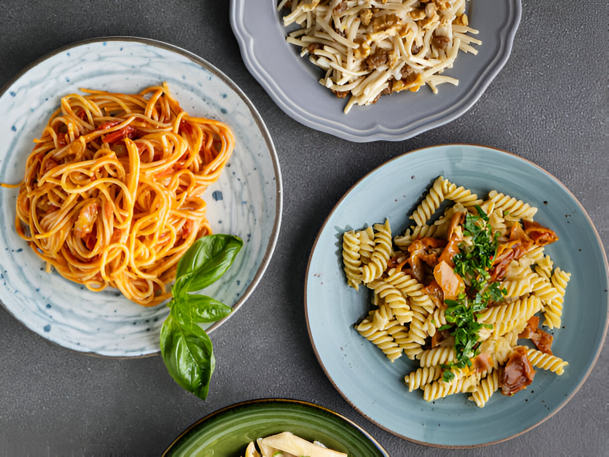Delicious variety of pasta dishes arranged on plates, including spaghetti with tomato sauce, fusilli with meat and herbs, and another pasta creation, set against a dark background.