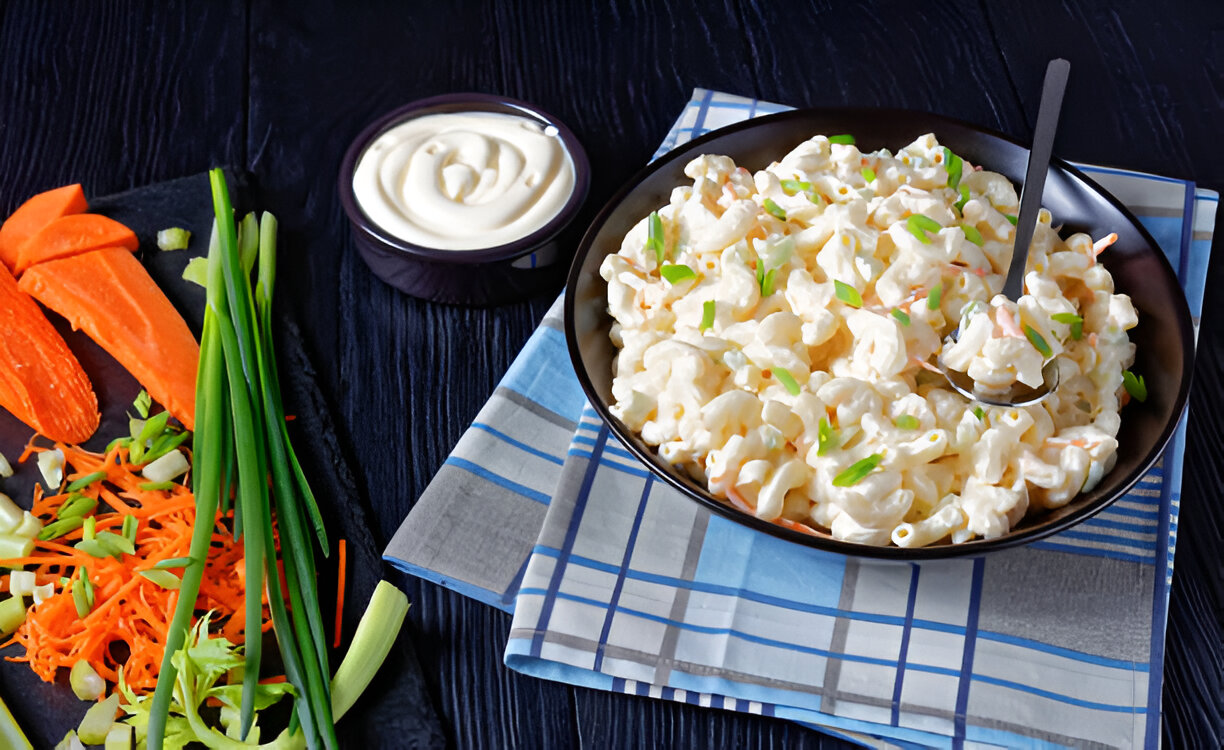 A bowl of creamy pasta salad garnished with green onions, alongside a platter of fresh cut vegetables including carrots, celery, and onions, with a small bowl of mayonnaise.