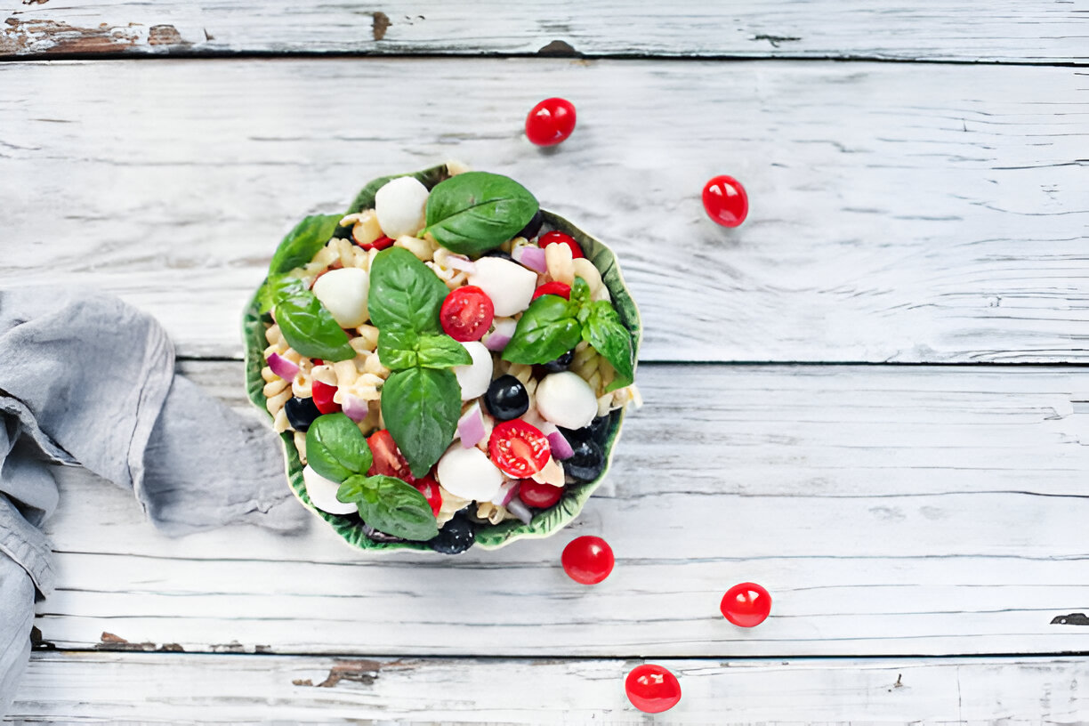 A vibrant pasta salad with cherry tomatoes, mozzarella balls, black olives, and fresh basil placed in a decorative bowl on a wooden surface.