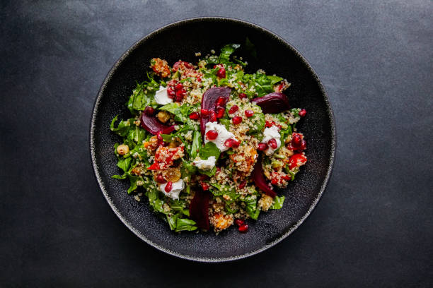 A vibrant salad featuring quinoa, greens, beets, pomegranate seeds, and a creamy dressing, served in a black bowl on a dark surface.
