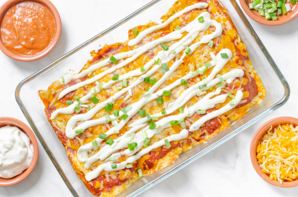 A glass dish filled with cheesy enchiladas topped with sour cream and green onions, surrounded by small bowls of salsa and cheese.