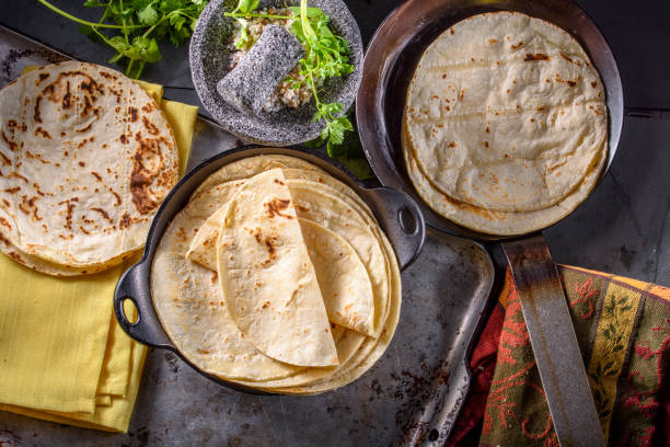 Freshly made tortillas stacked in a bowl with additional tortillas in a frying pan, garnished with herbs and accompanying dipping sauce.
