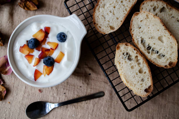 A bowl of yogurt topped with chopped peaches and blueberries, next to slices of bread on a wire rack.