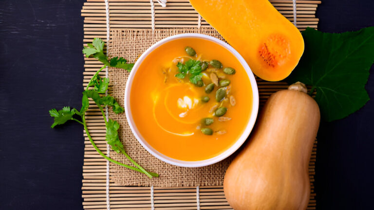 A bowl of creamy butternut squash soup topped with seeds and fresh herbs, placed on a bamboo mat next to cut butternut squash.
