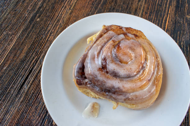 A delicious cinnamon roll topped with glaze on a white plate placed on a rustic wooden table.