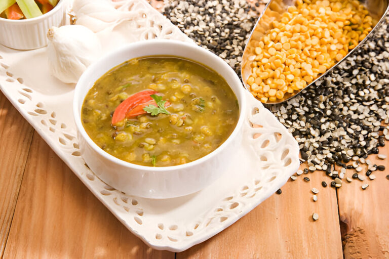 A bowl of green lentil soup garnished with tomato and coriander served on a decorative platter with raw garlic and mixed lentils in the background.