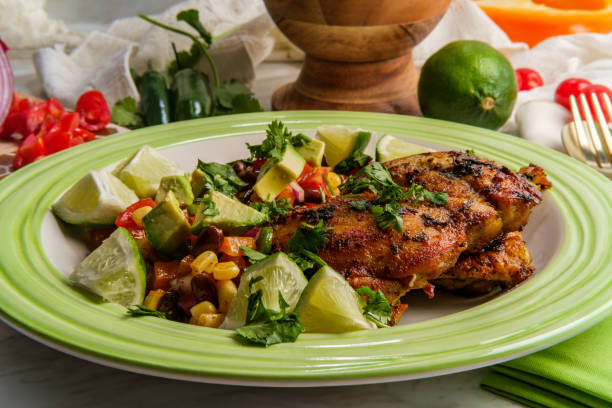 A vibrant plate of grilled chicken served with fresh avocado, lime wedges, and a colorful salad featuring tomatoes, corn, and beans.