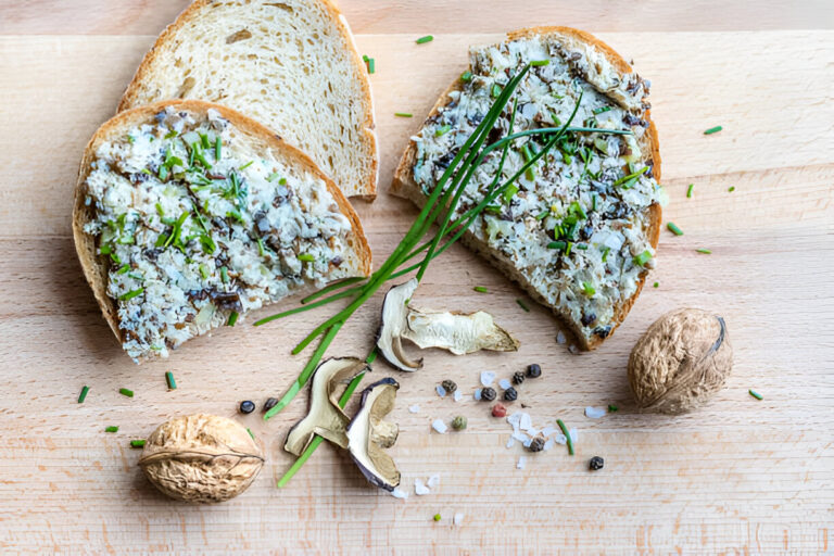 Two slices of bread topped with a creamy spread, garnished with chives, placed on a wooden surface alongside walnuts and dried mushrooms.