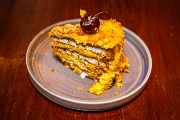 A slice of layered cake with yellow frosting and a cherry on top, presented on a round plate.