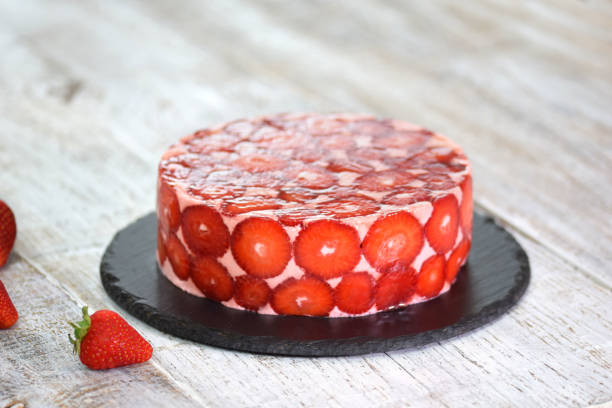 A round strawberry cake with a glossy red glaze, decorated with fresh strawberries on a black slate plate.