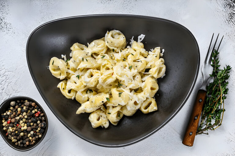 A bowl of creamy pasta with herbs and cheese, accompanied by a small bowl of mixed peppercorns and a fork.