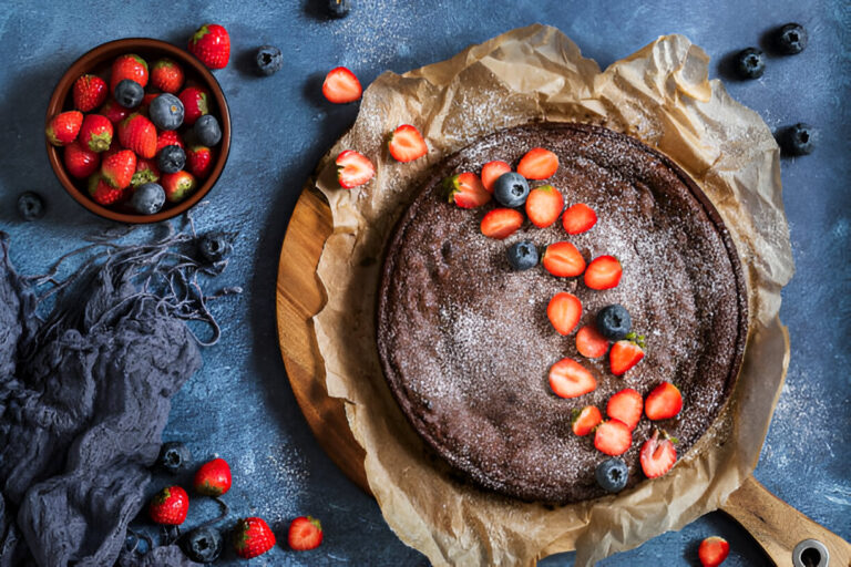 A delicious chocolate cake decorated with fresh strawberries and blueberries, placed on a wooden board with parchment paper, against a blue background.