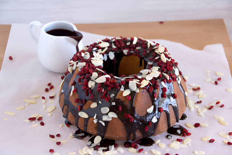 Bundt cake topped with chocolate glaze, dried raspberries, and sliced almonds, with a small white jug of chocolate sauce in the background.