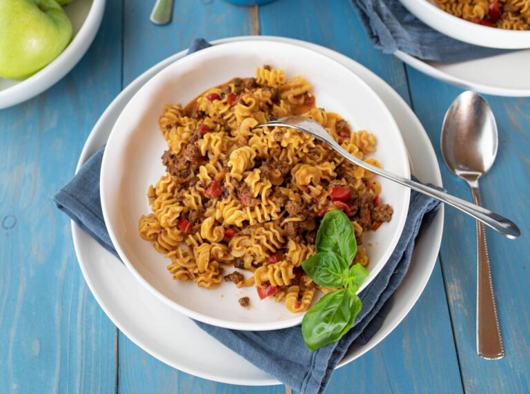 A bowl of pasta with ground meat and diced tomatoes, garnished with fresh basil leaves, on a blue wooden table.