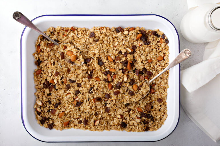 A rectangular dish filled with granola, featuring nuts and chocolate chips, with two serving spoons resting inside.