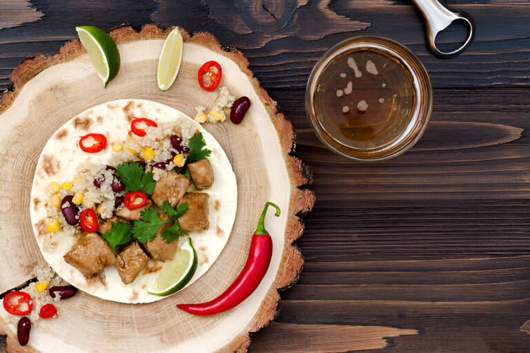 A delicious taco filled with meat, quinoa, corn, and sliced red chilies, served on a wooden platter with lime wedges and a drink