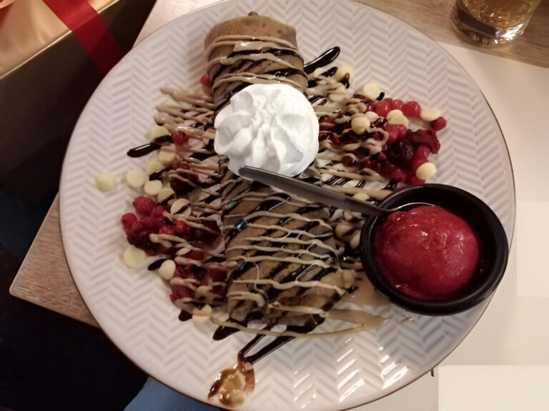 A delicious dessert plate featuring a chocolate-filled crepe topped with whipped cream, raspberry coulis, and drizzled with chocolate and caramel sauces, accompanied by a scoop of raspberry sorbet.