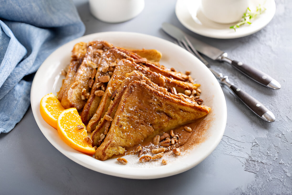 A plate of French toast topped with crushed peanuts and powdered sugar, garnished with orange slices, served on a light gray surface.
