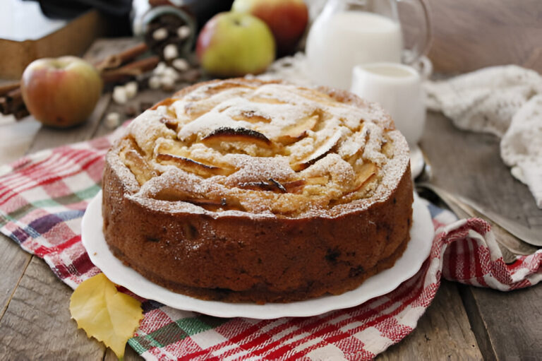 A freshly baked apple cake dusted with powdered sugar, placed on a white plate on a wooden table with apples and milk in the background.