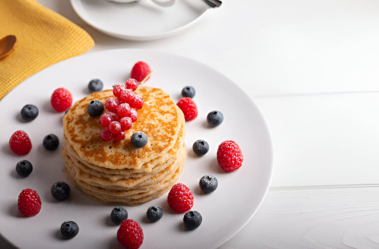 A stack of pancakes topped with red currants and surrounded by fresh raspberries and blueberries on a white plate.