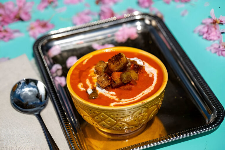 A vibrant bowl of soup garnished with croutons, placed on a silver tray with pink flower petals scattered around.