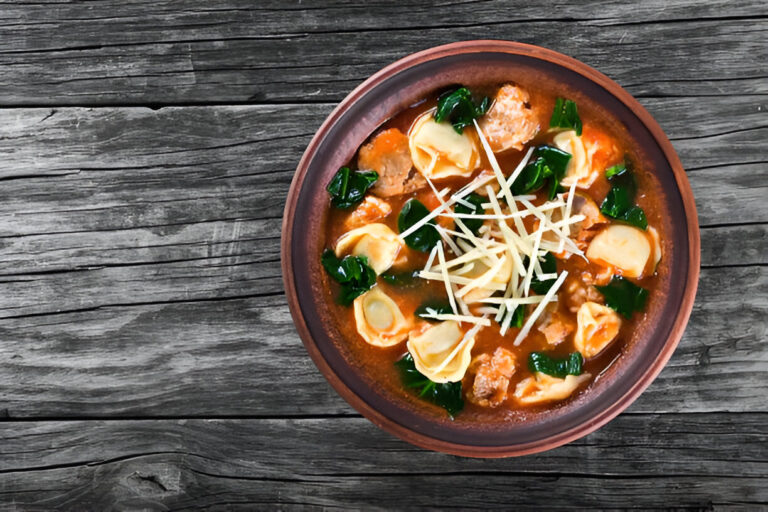 A bowl of soup with tortellini, leafy greens, and garnished with sliced ginger on a rustic wooden surface.