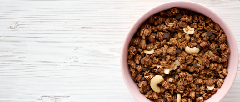 A bowl of granola mixed with nuts and chocolate chips on a white wooden table.