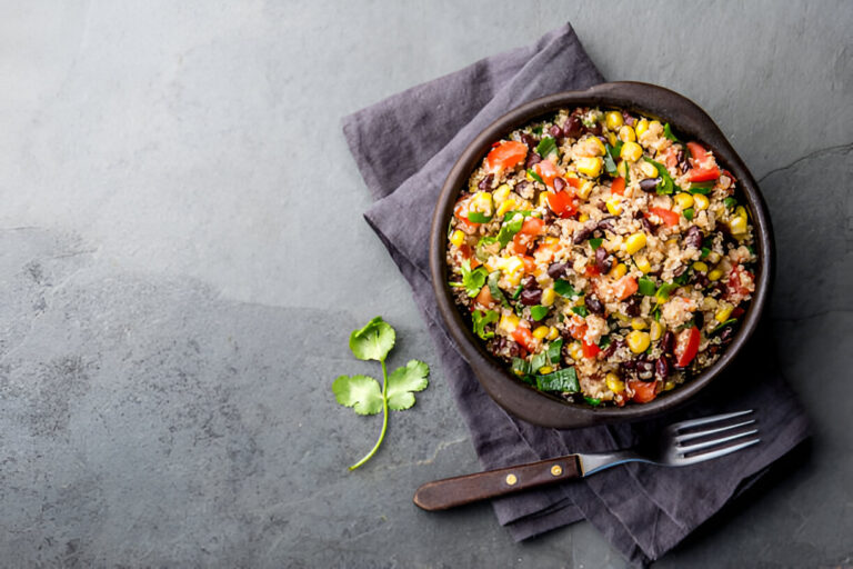A bowl of quinoa salad filled with black beans, corn, diced tomatoes, and fresh herbs on a gray stone surface with a fork and napkin.