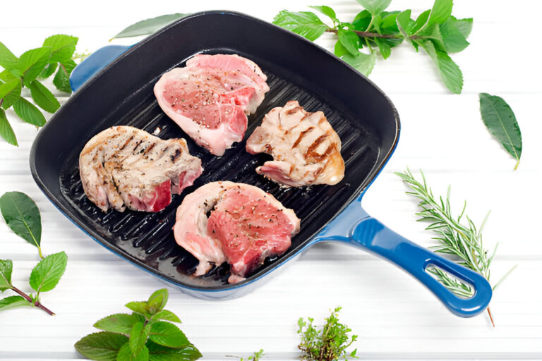 Grilled meat chops on a blue grill pan surrounded by fresh herbs