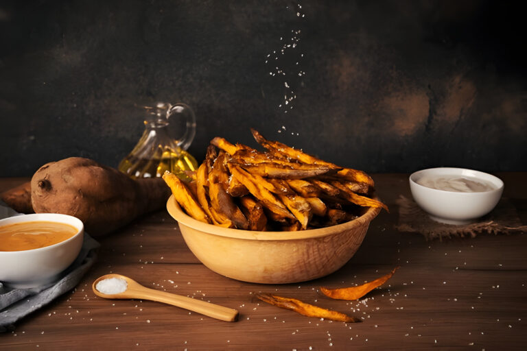 A wooden bowl filled with crispy sweet potato fries, sprinkled with salt, alongside a spoon of salt, a bowl of dipping sauce, and a sweet potato, with olive oil in the background.