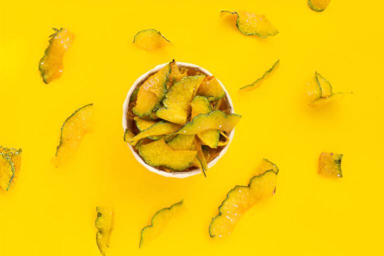 A bowl filled with yellow pumpkin chips surrounded by scattered pumpkin peel on a vibrant yellow background.