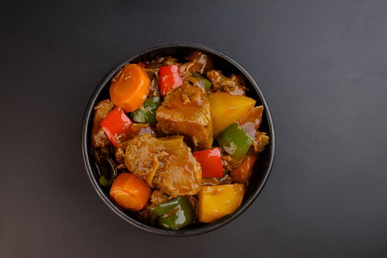 A bowl of colorful stir-fried vegetables and meat, including red, yellow, and green peppers, carrots, and sauce, served on a black surface.