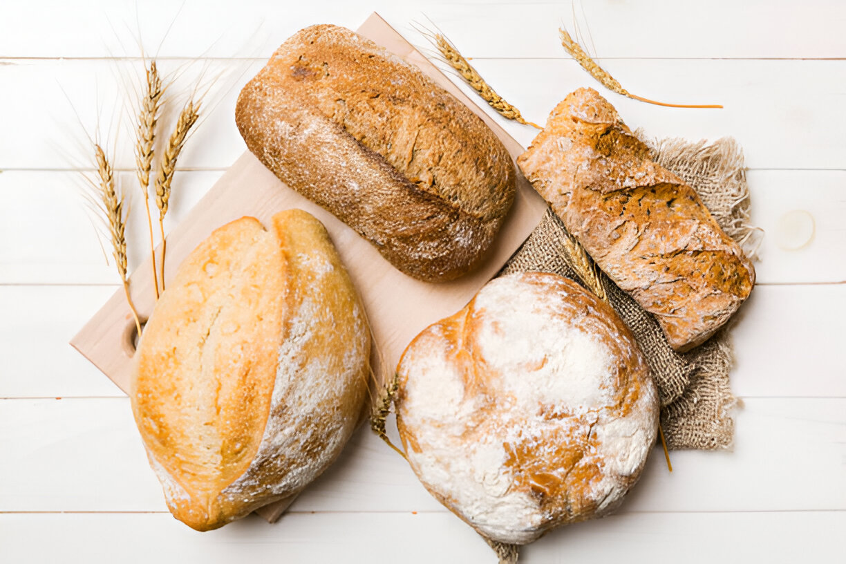 An assortment of fresh bread loaves on a wooden board with decorative wheat stalks in the background.