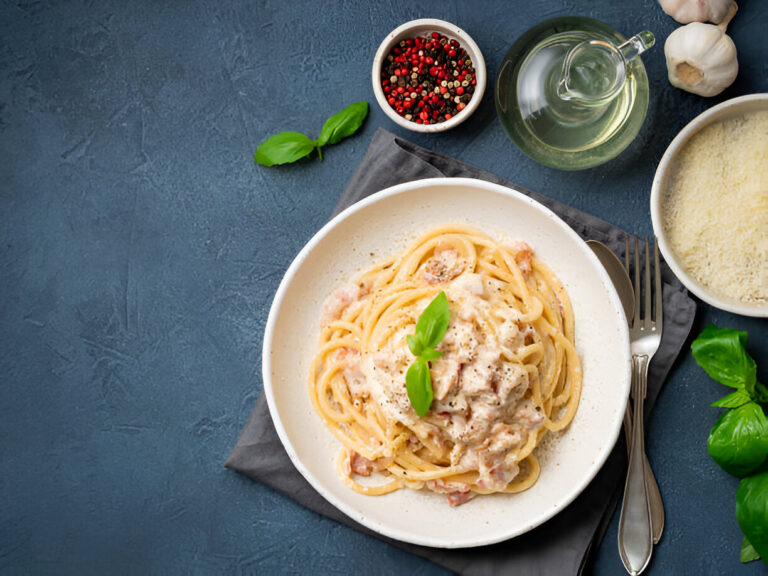 A delicious serving of creamy pasta with basil on a plate beside peppercorns, olive oil, garlic, and grated cheese.