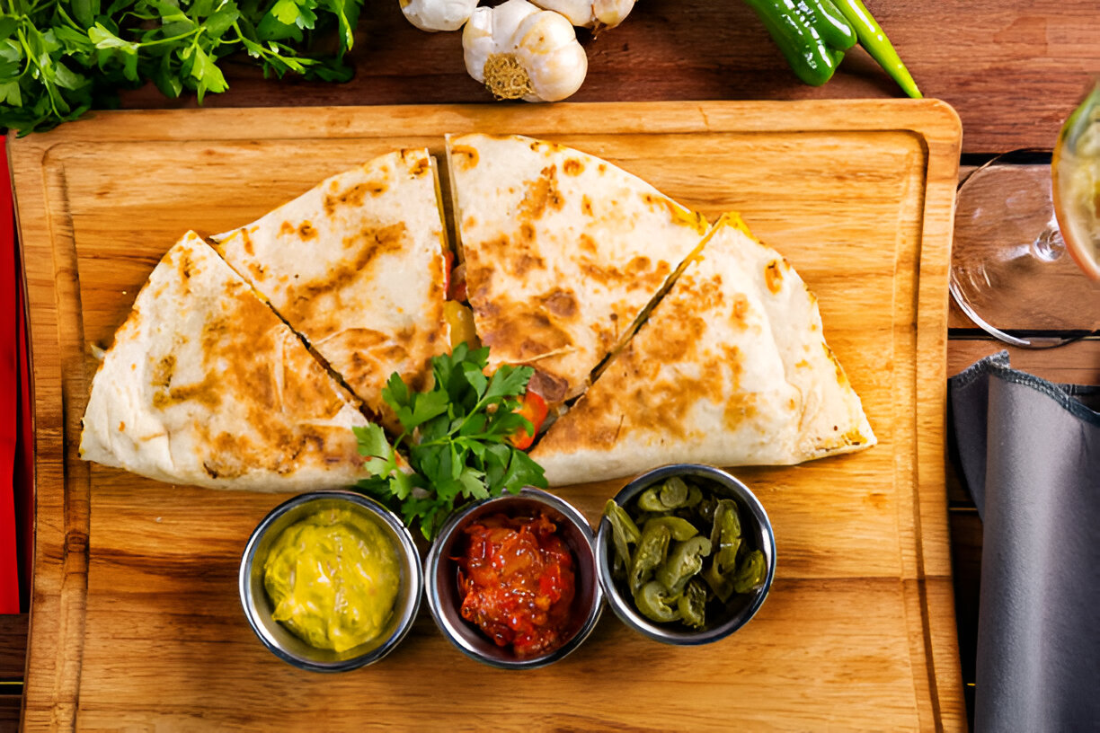 A delicious quesadilla cut into slices, placed on a wooden serving board, accompanied by bowls of salsa, guacamole, and jalapenos.