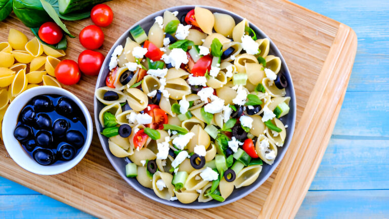 A colorful pasta salad with shell-shaped pasta, cherry tomatoes, green peppers, olives, and feta cheese on a wooden board.