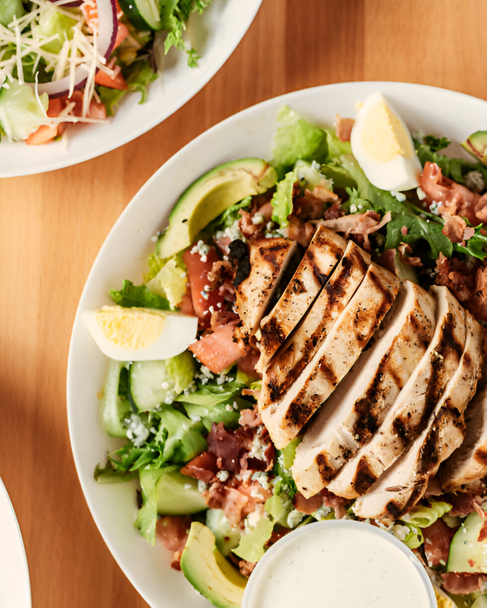 Grilled chicken salad with sliced chicken breast, avocado, and hard-boiled egg on a bed of greens, accompanied by a creamy dressing.