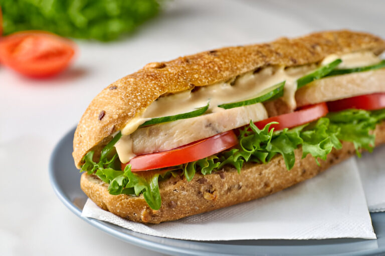 A close-up of a healthy sandwich with whole grain bread, lettuce, sliced tomatoes, cucumbers, and a creamy sauce.