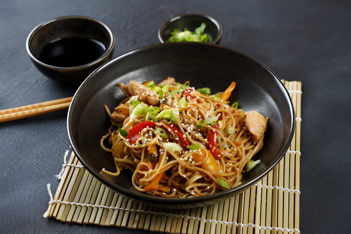 A bowl of stir-fried noodles with vegetables and chicken, garnished with green onions and sesame seeds, served with a side of soy sauce.