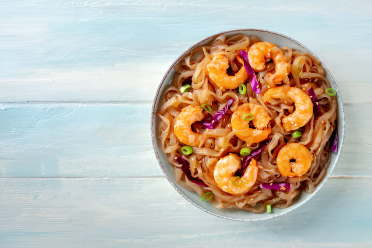 A bowl of shrimp noodles with purple cabbage and green onions on a light blue background.